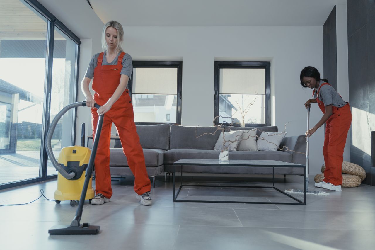 why-choose-us Two women in red overalls vacuuming and sweeping a modern living room.